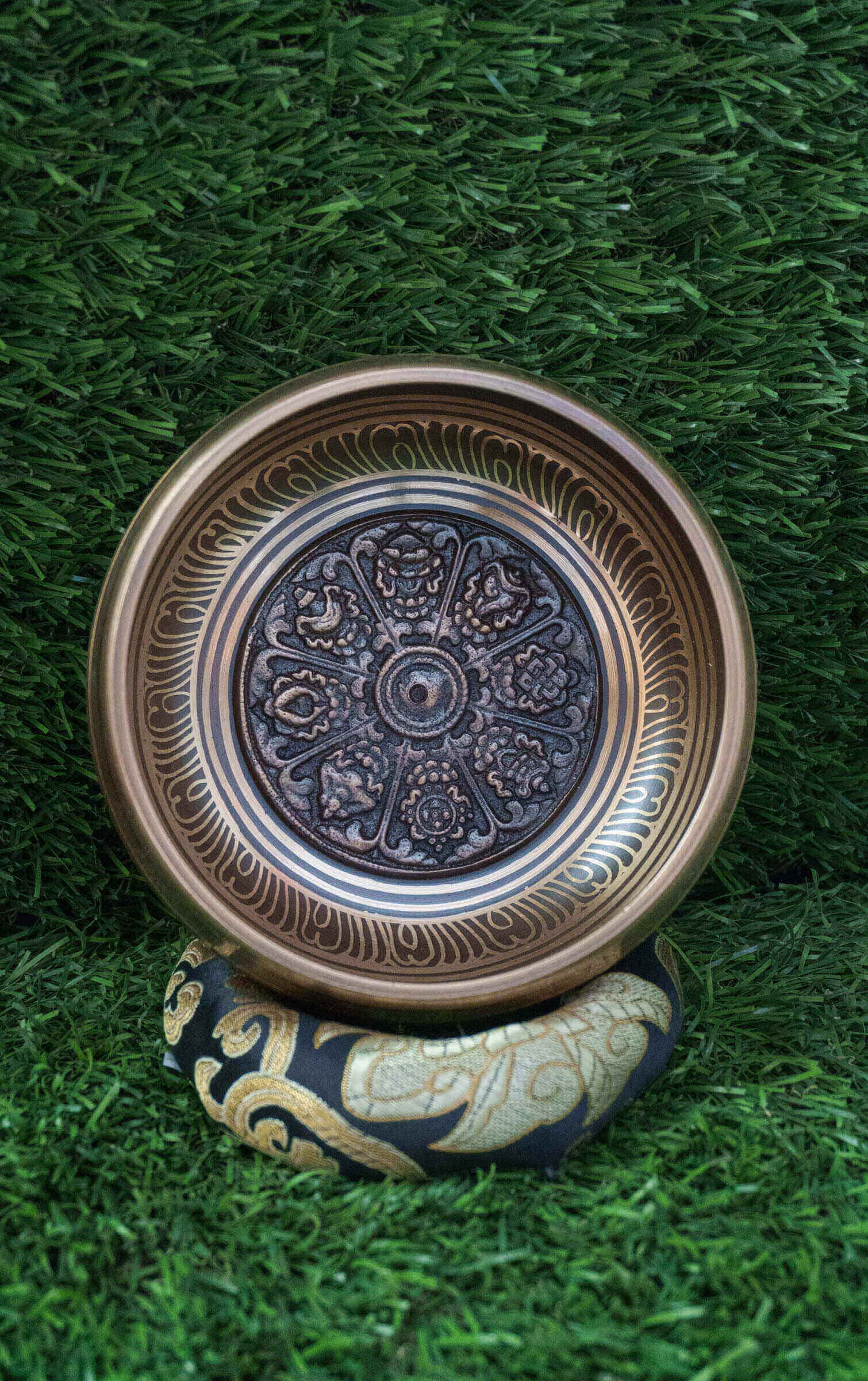 Top-down view of a bronze Tibetan singing bowl featuring an engraved Ashtamangala (Eight Auspicious Symbols) mandala design on its interior, resting on a black and gold silk cushion over green grass.
