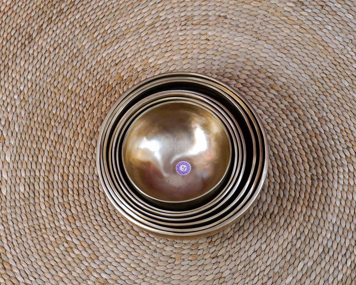 Seven handcrafted singing bowls nested together, showing their graduated sizes, smooth metal surfaces, and warm bronze-gold finish.
