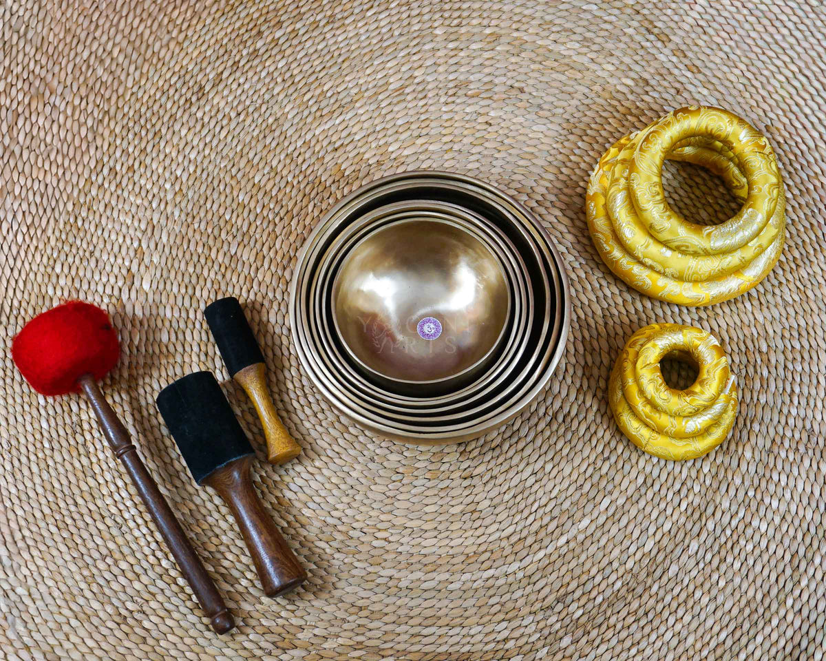 Seven chakra-inspired singing bowls arranged with wooden mallets and decorative golden cushions for meditation and sound practice.