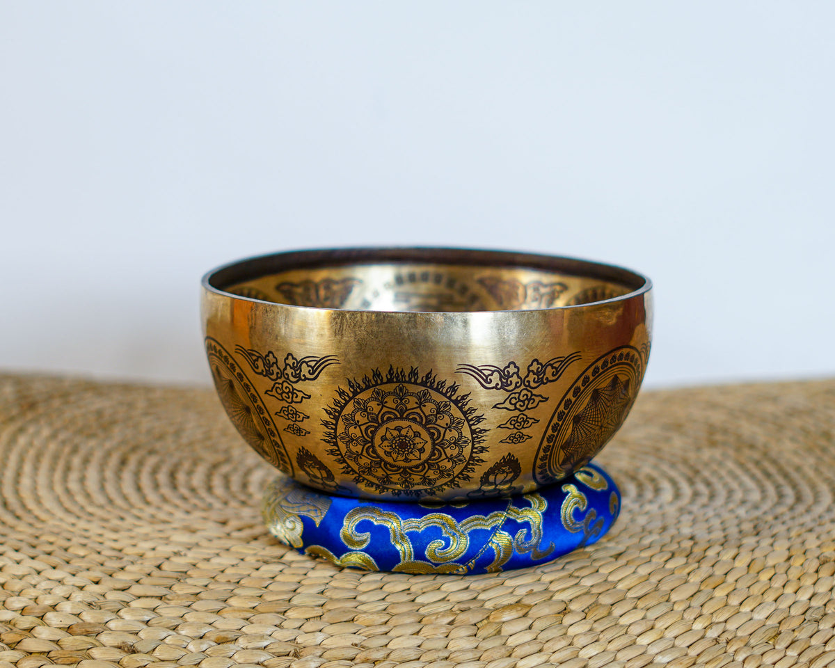 Golden floral mandala singing bowl with detailed ornamental engravings displayed on a blue brocade cushion.