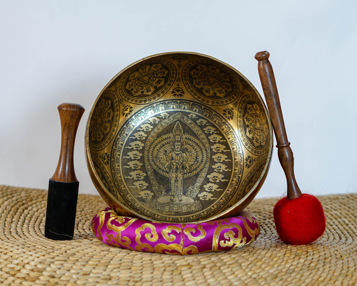Avalokiteshvara singing bowl displayed upright with a wooden mallet and red felt mallet, showing intricate Buddhist engravings.