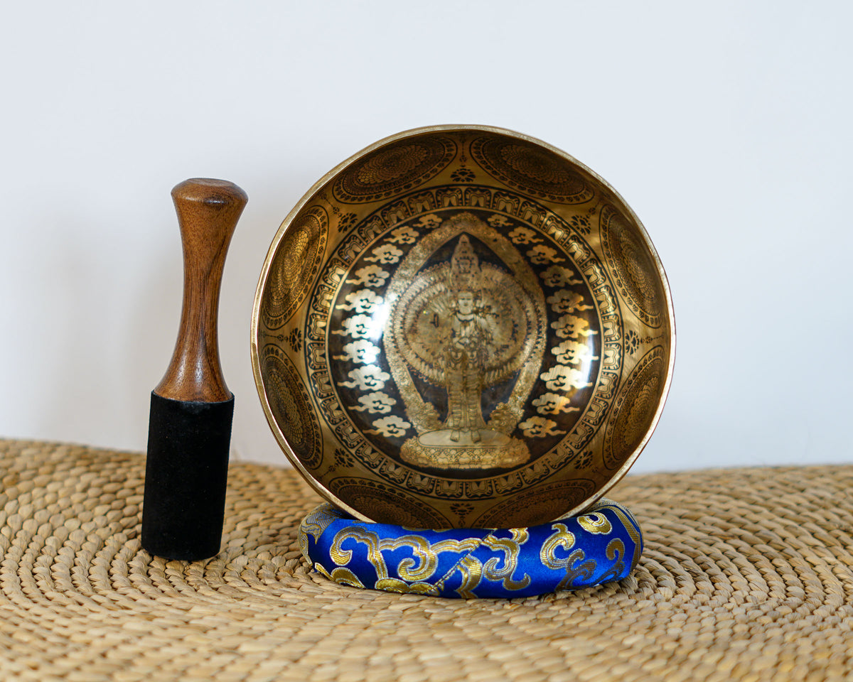 Thousand Armed Avalokiteshvara singing bowl shown upright with a wooden mallet and blue cushion, displaying the intricate Buddhist engraving.