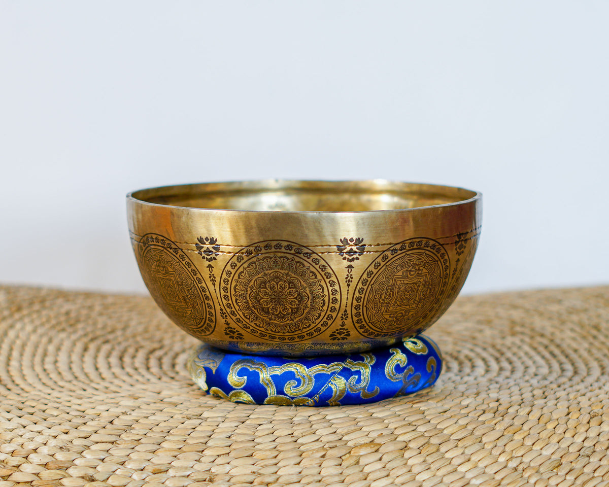 Kalchakra Yogini Singing Bowl resting on a blue brocade cushion, displaying intricate mandala engravings on its exterior.
