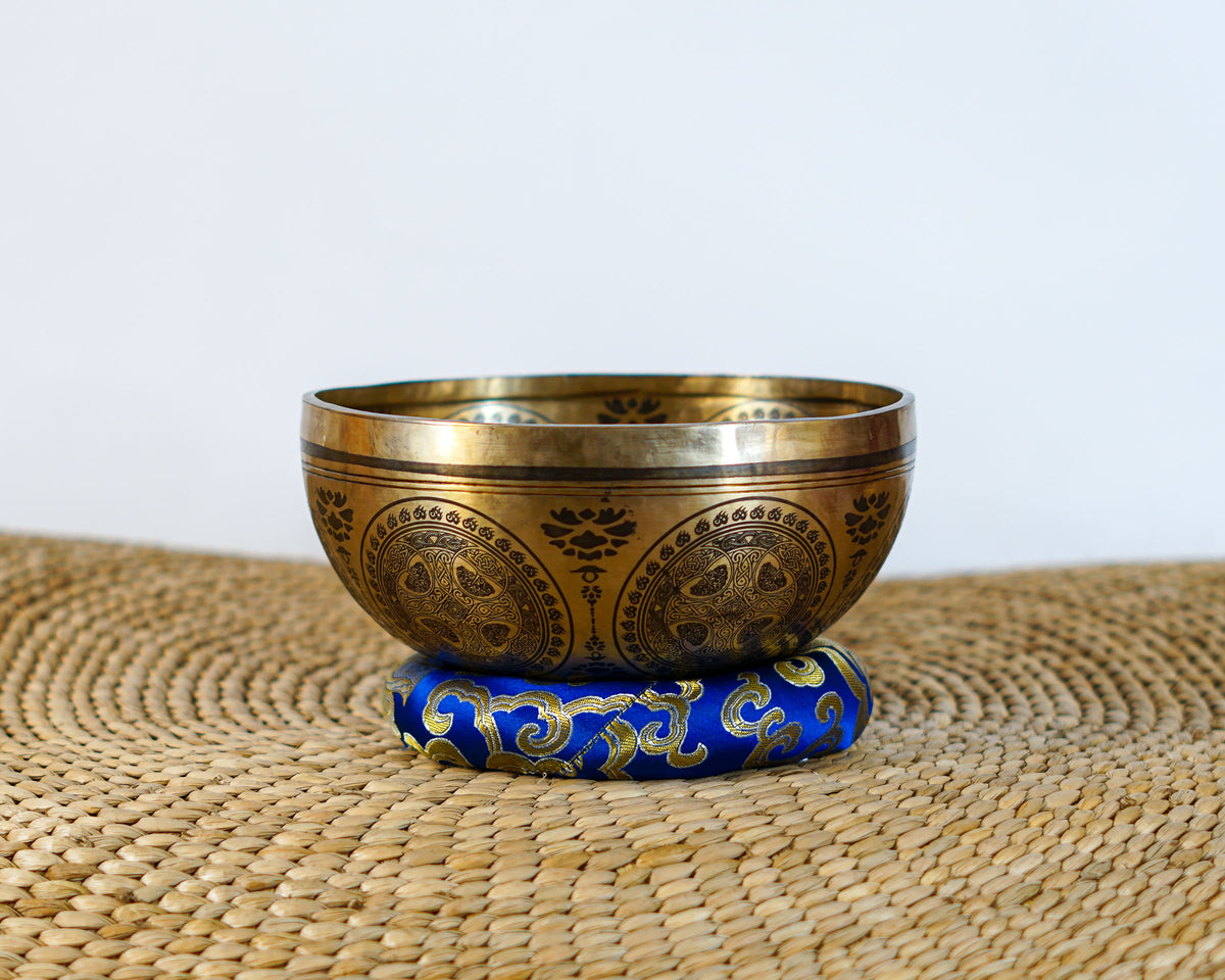 Golden Seven Chakra singing bowl displayed on a blue brocade cushion with decorative engraved patterns.