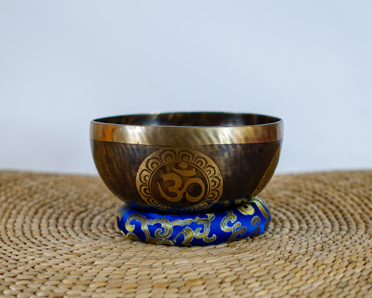 Handmade Hindu Om singing bowl with antique gold rim and engraved Om motif displayed on a blue brocade cushion.