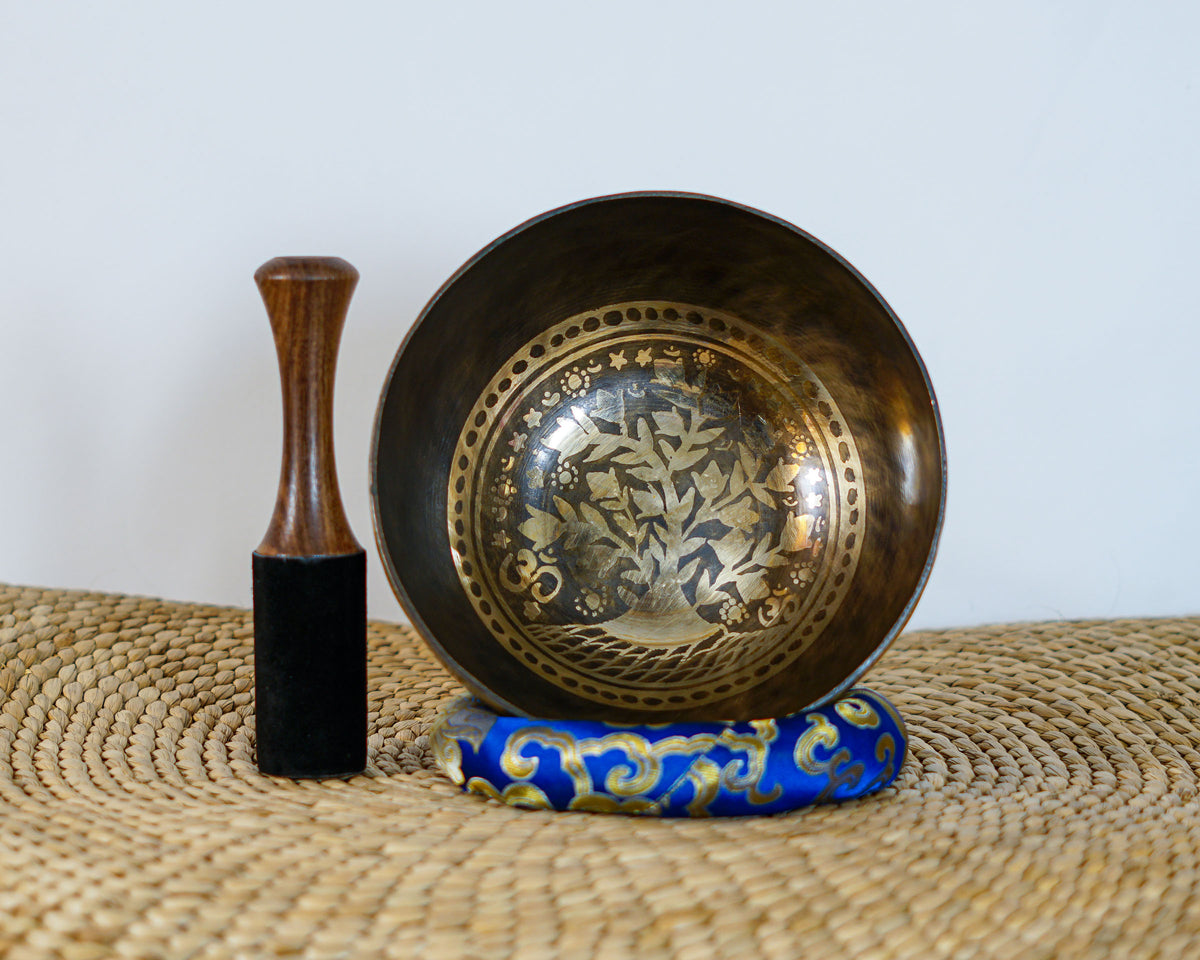 Tree of Life engraved singing bowl displayed upright with a wooden mallet and blue brocade cushion on a woven mat.