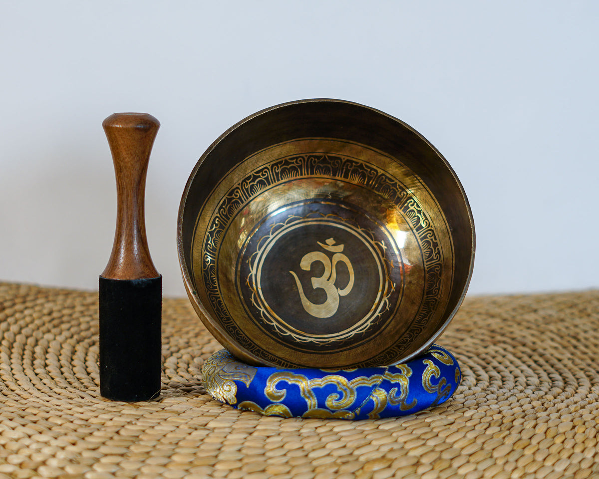 Hindu Om singing bowl standing upright with a wooden mallet and blue brocade cushion, showing the central Om engraving.