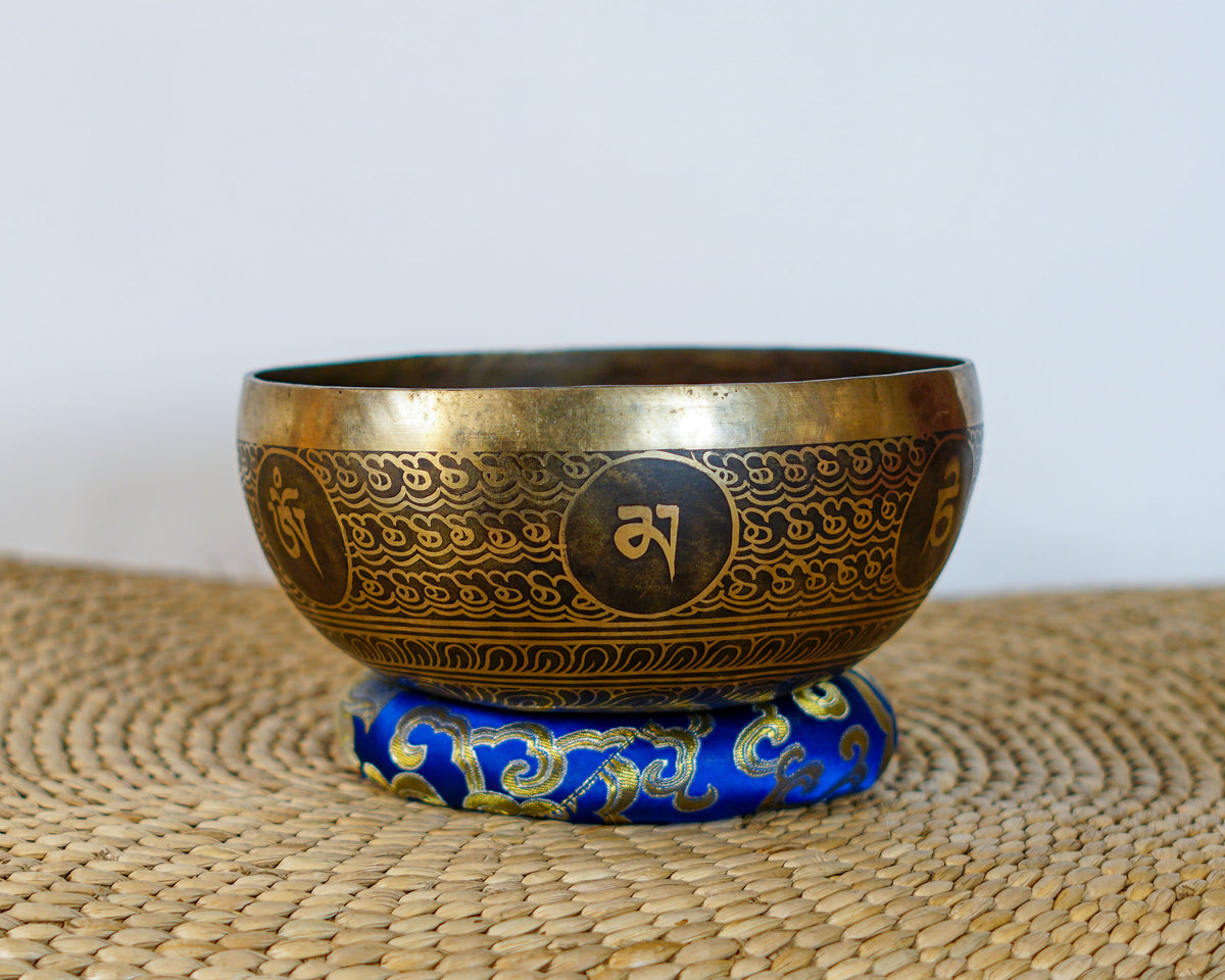 Lotus engraved singing bowl with golden exterior patterns resting on a blue brocade cushion.