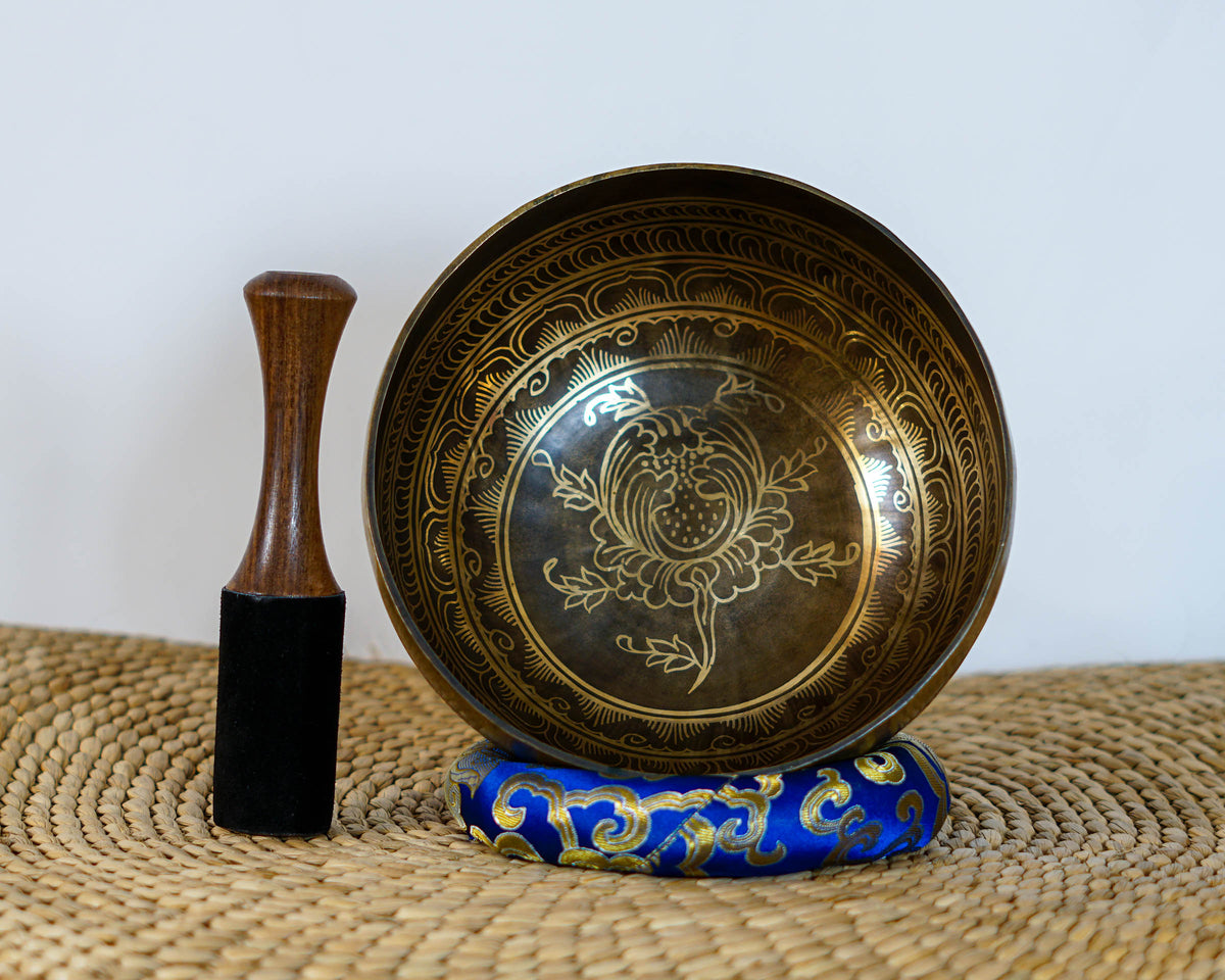 Engraved lotus singing bowl displayed upright with a wooden mallet and blue brocade cushion, showing the intricate golden lotus design inside.