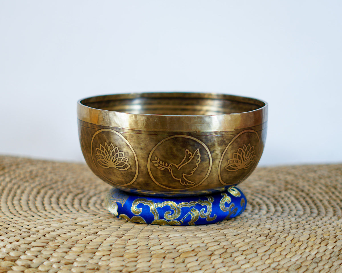 Golden bronze Dove Tibetan singing bowl featuring engraved dove and lotus flower designs on a blue brocade cushion.