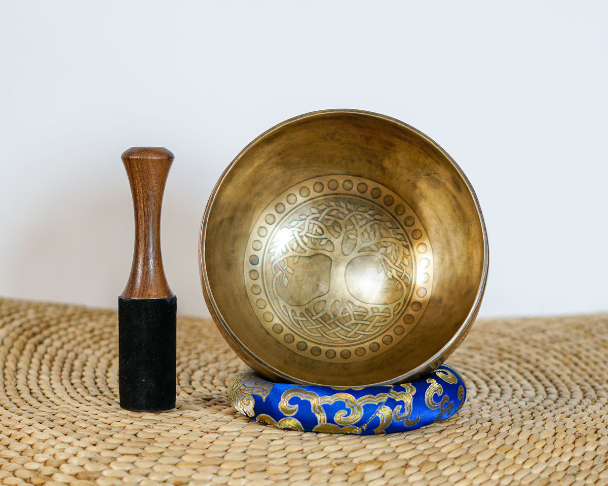 Tree of Life engraved Tibetan singing bowl displayed upright with a wooden striker and blue brocade cushion, showing the detailed interior artwork.