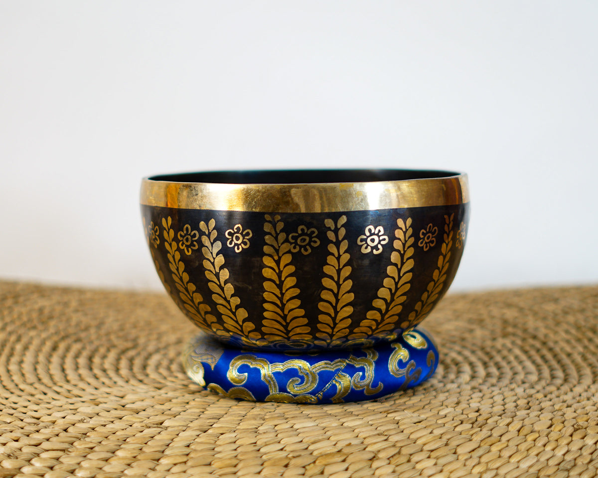 Dark bronze Tibetan singing bowl with golden Tree of Life inspired leaf and floral engravings displayed on a blue brocade cushion.