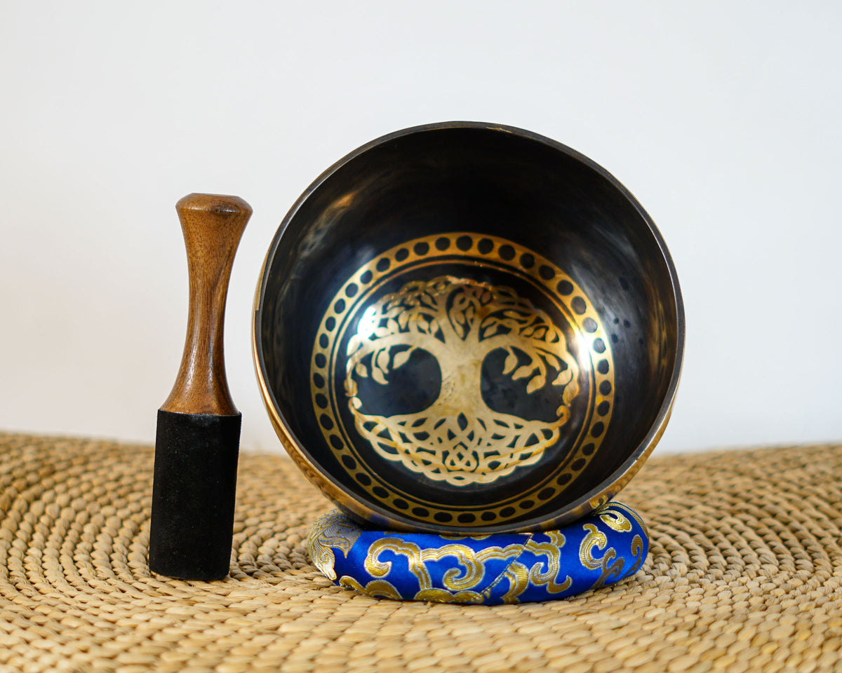 Tree of Life Tibetan singing bowl with black and gold interior engraving, photographed upright with a wooden striker and blue brocade cushion.