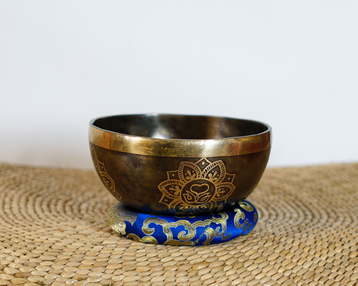 Dark antique bronze Tibetan singing bowl with a golden Swan Symbol of Life engraving on the exterior, displayed on a blue brocade cushion.