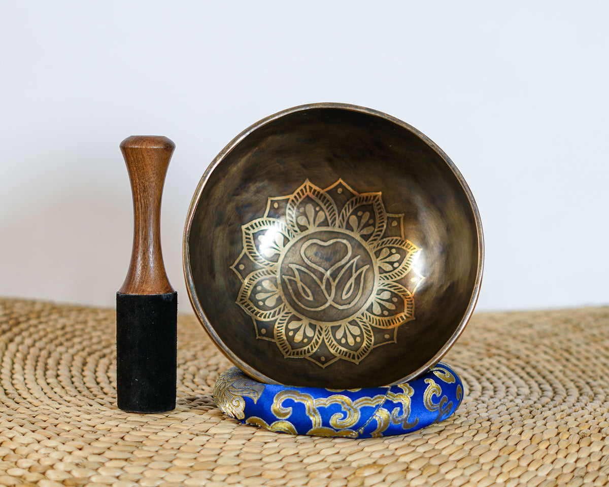 Engraved Tibetan singing bowl featuring the Swan Symbol of Life and decorative golden floral patterns, photographed upright with a wooden striker and blue brocade cushion.