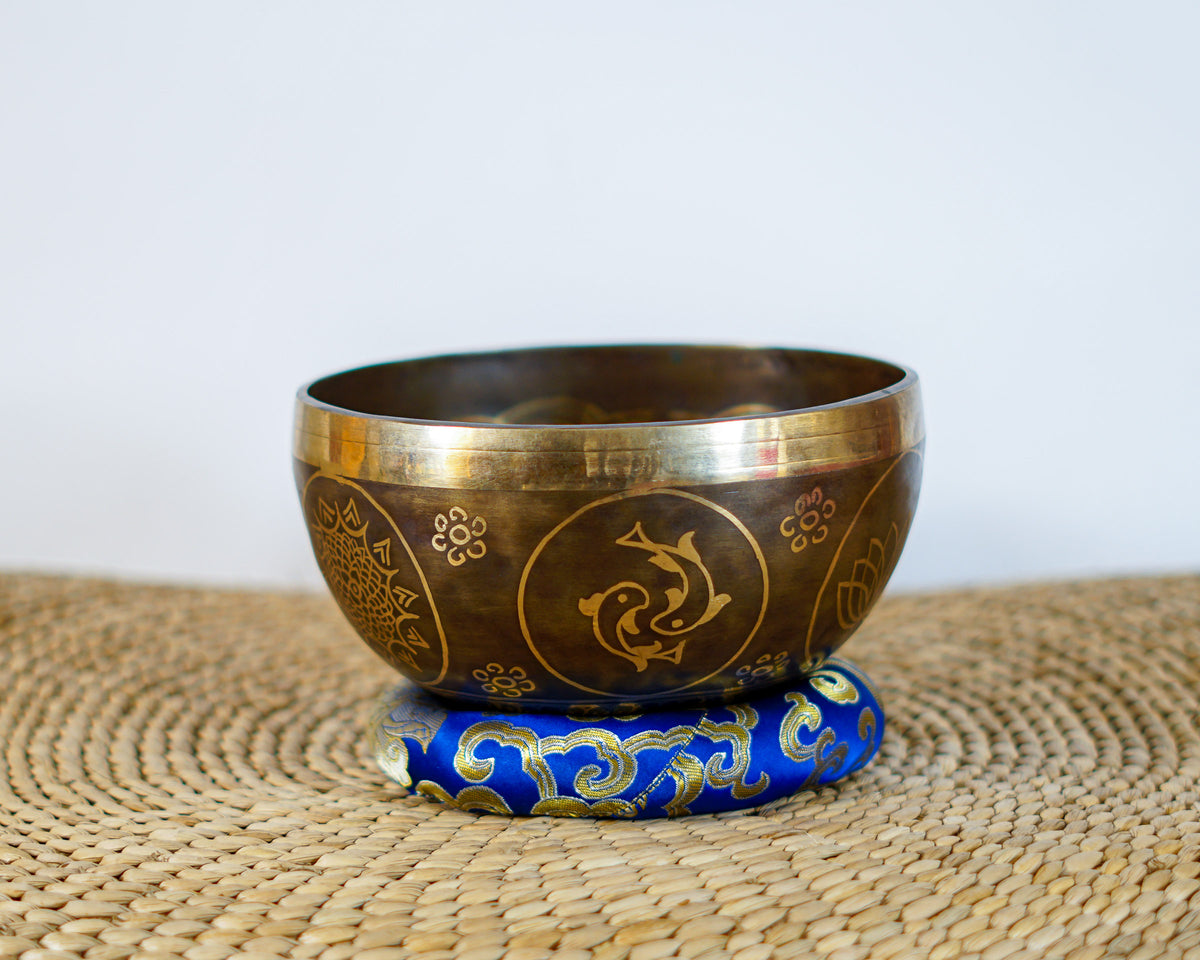 Side exterior view of a dark antique bronze Tibetan singing bowl with golden engraved fish motifs, resting on a blue brocade cushion.