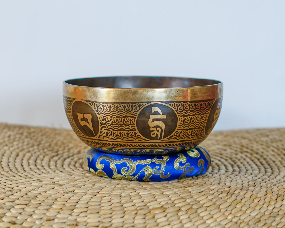 Golden Tibetan singing bowl with Tibetan script and decorative engraved borders displayed on a blue brocade cushion.
