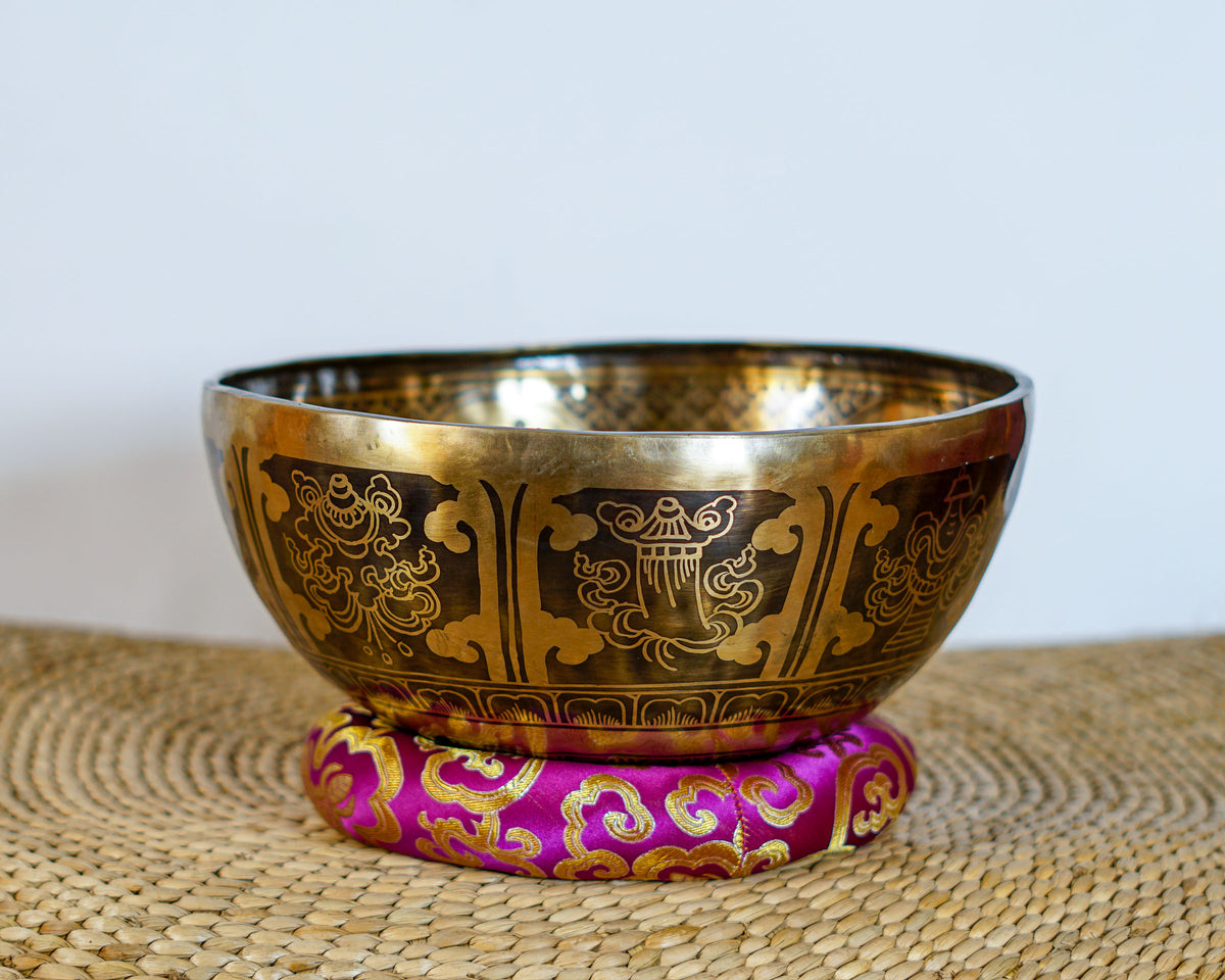 Golden Tibetan singing bowl featuring engraved Buddhist symbols and decorative patterns, displayed on a pink brocade cushion.