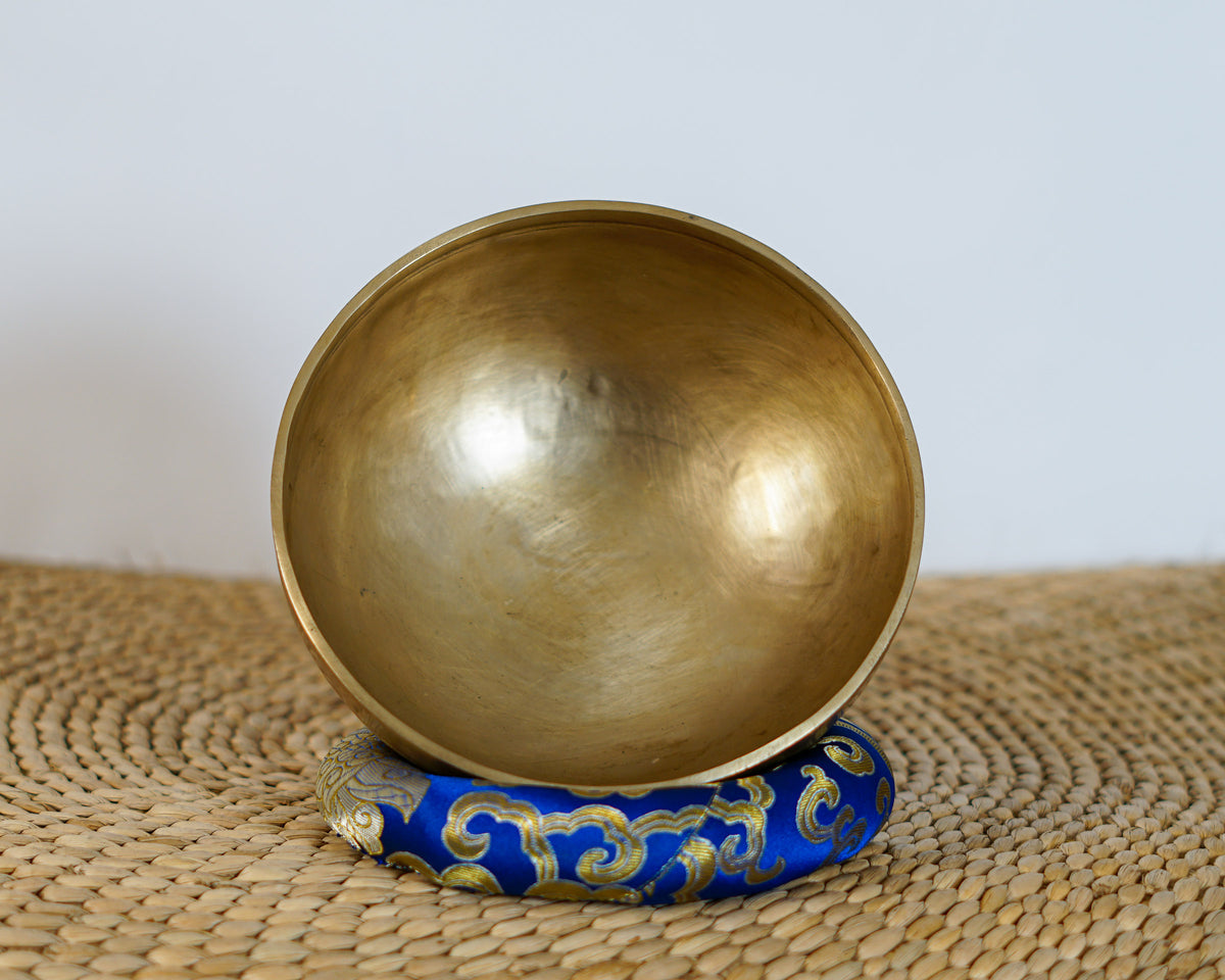 Side profile view of a small warm golden hand-hammered bronze singing bowl with double rim lines, displayed on a blue brocade cushion.