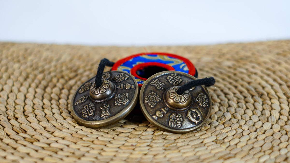 Pair of Tibetan Buddhist tingsha cymbals with eight auspicious symbols displayed beside a colorful brocade case.