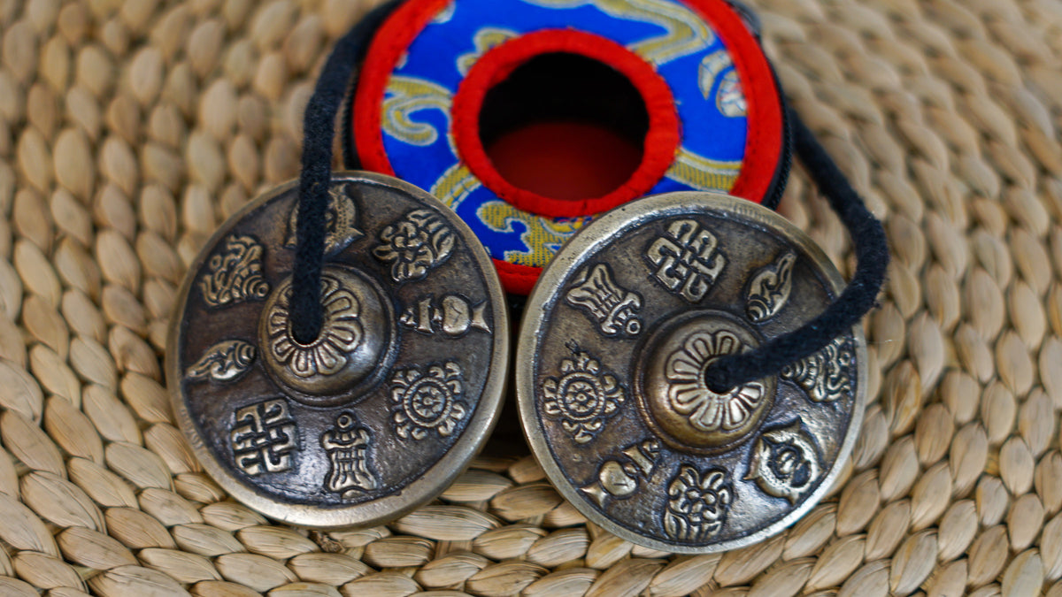 Close up of Tibetan tingsha cymbals showing raised auspicious symbols, antique bronze tones, and black connecting cord.