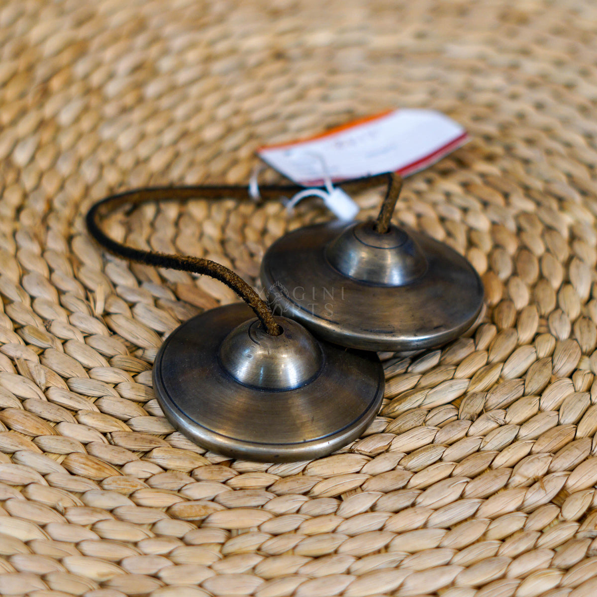 Traditional Tibetan Tingsha cymbals handcrafted from metal with a dark antique finish and connected by a cord, photographed on a natural woven surface.