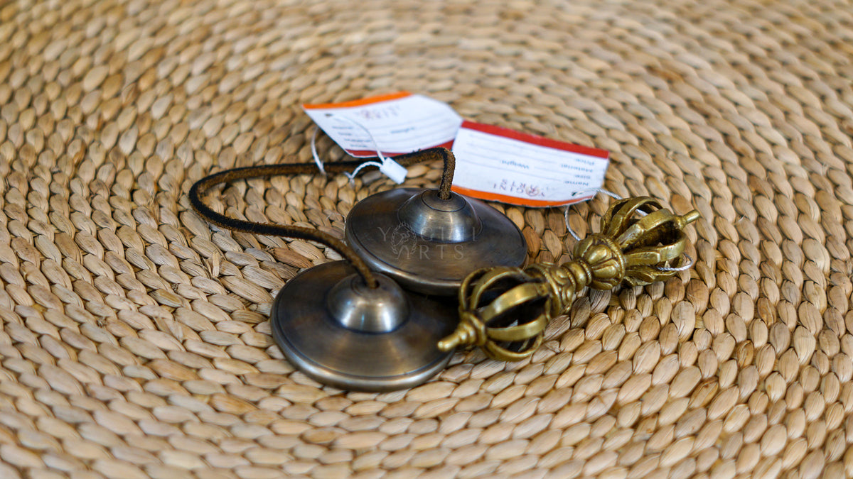 Traditional Tibetan Tingsha cymbals handcrafted from metal with a dark antique finish and connected by a cord, photographed on a natural woven surface.