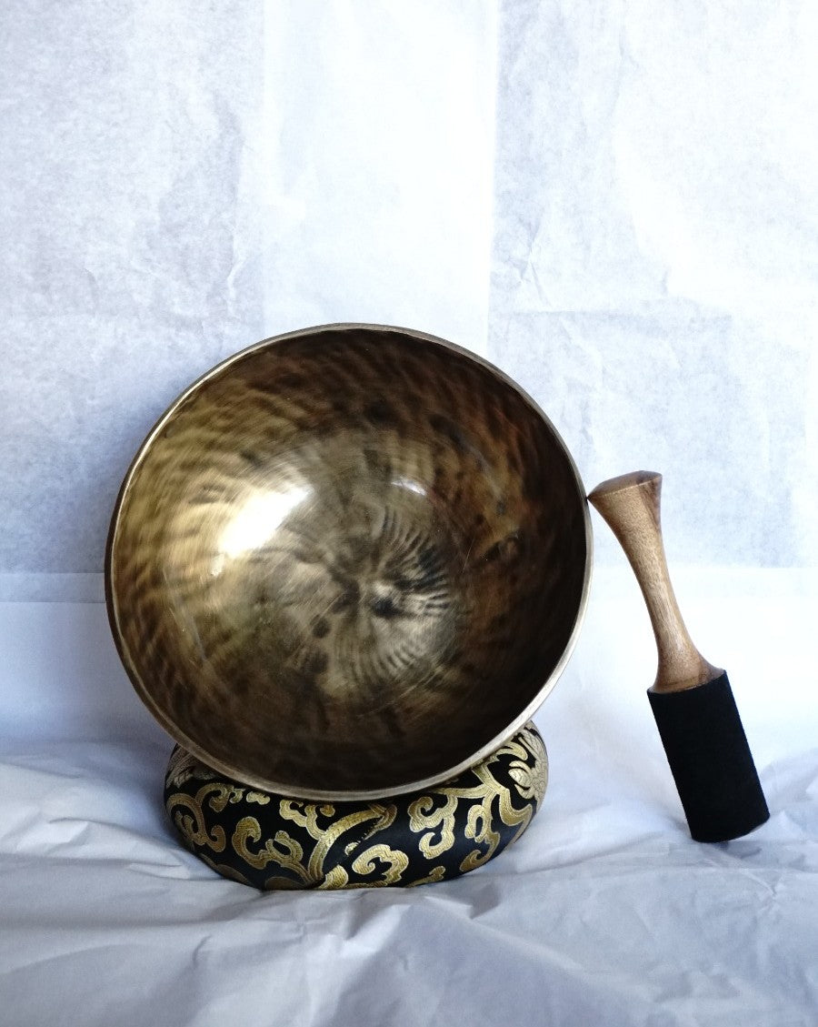 Hand-hammered brass singing bowl placed on a decorative brocade cushion with a wooden striker beside it, displayed against a white background.