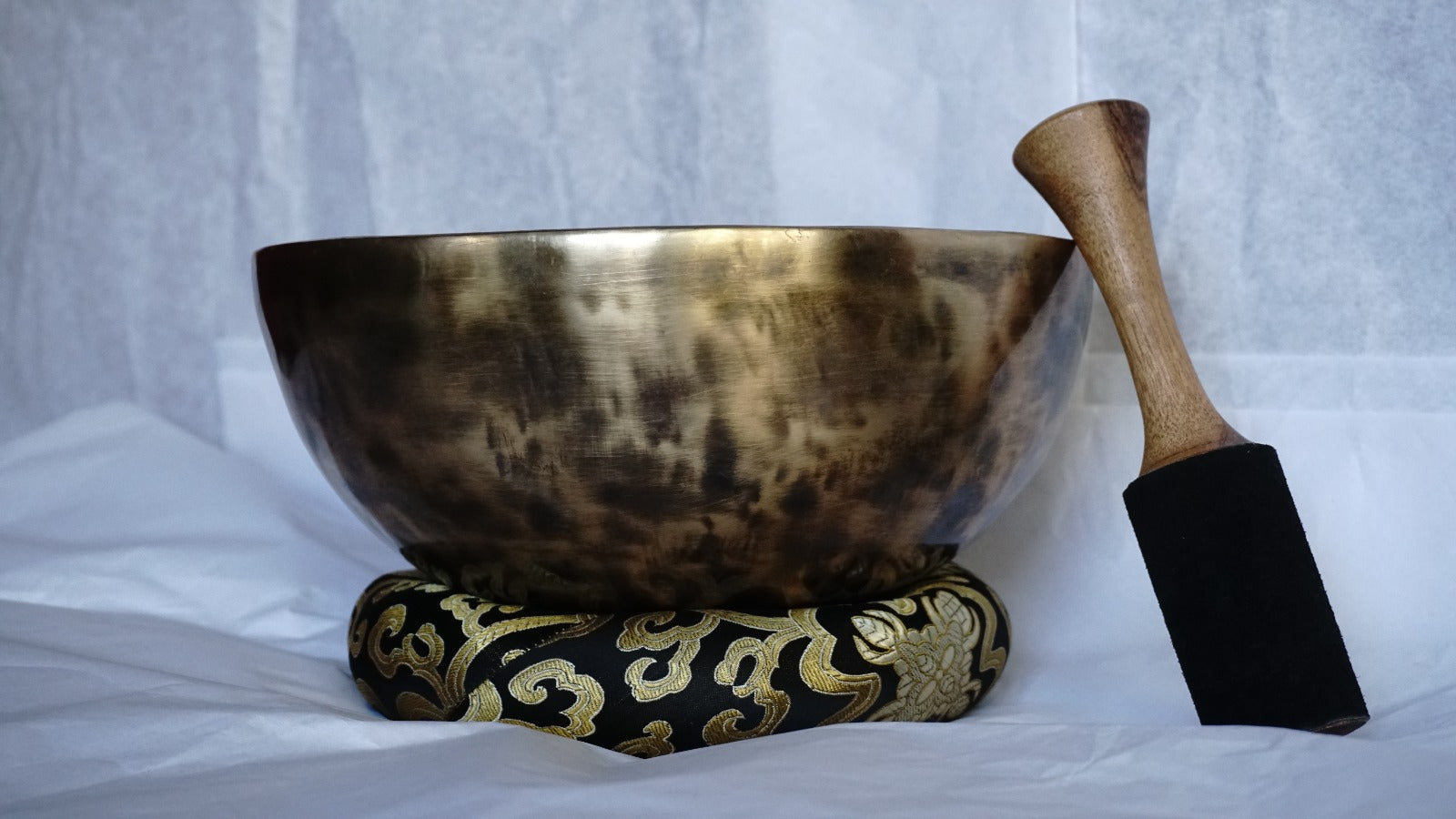 Hand-hammered brass singing bowl placed on a decorative brocade cushion with a wooden striker beside it, displayed against a white background.