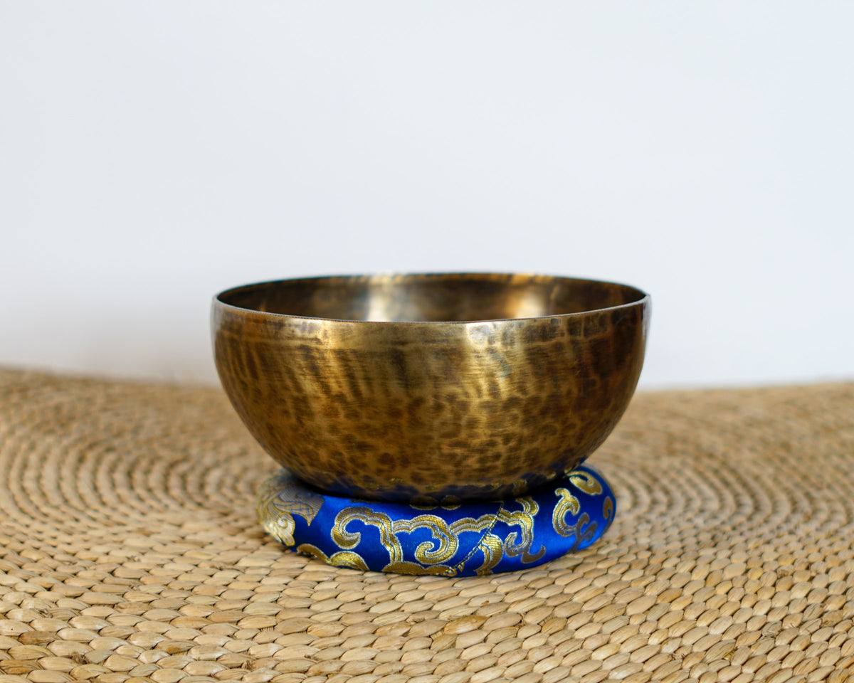 Side profile view of a small golden bronze Tiger style hand-hammered singing bowl with mottled surface patina, resting on a blue brocade cushion.