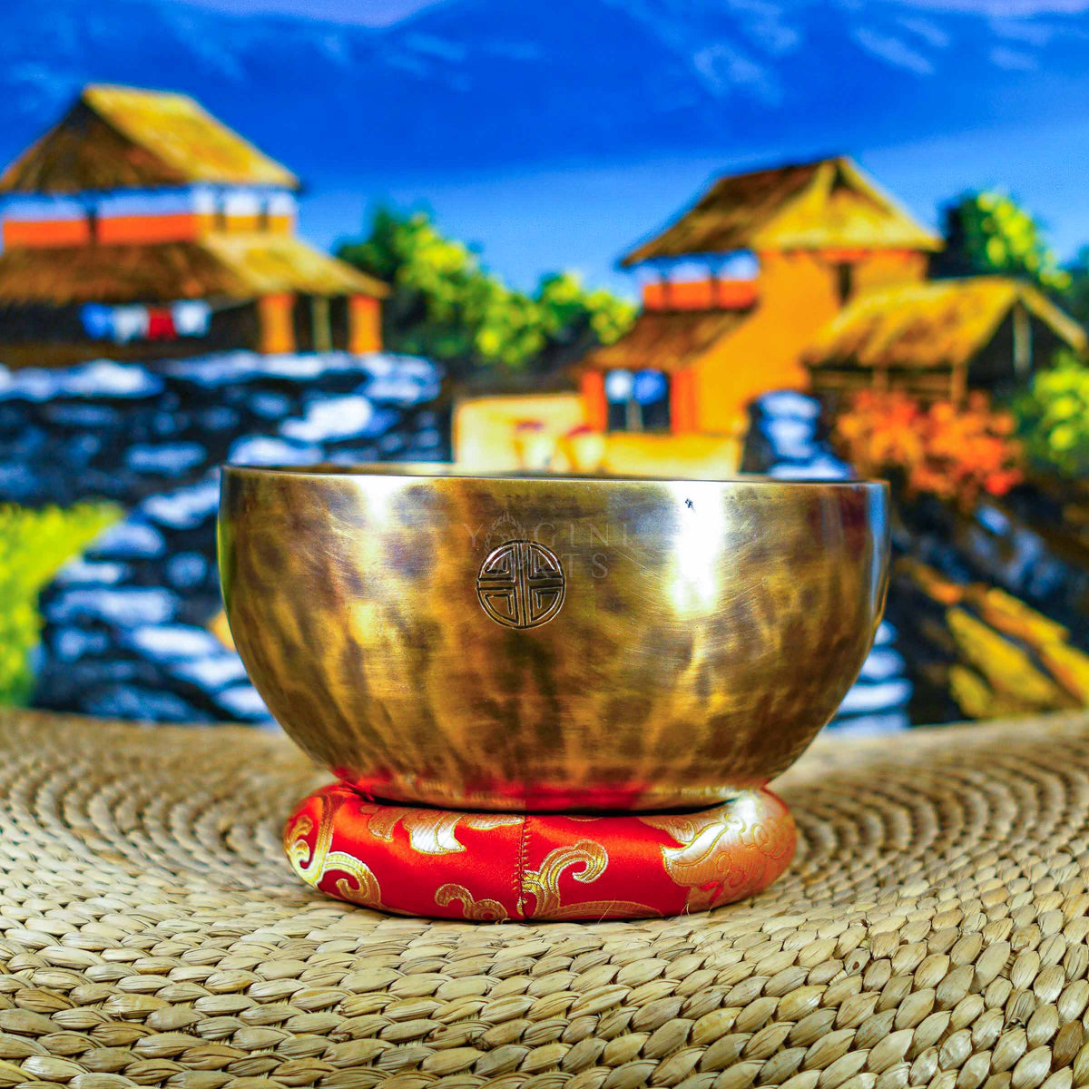 Handcrafted seven chakra singing bowl set with a brass-toned bowl, Om symbol, red cushion and wooden-handled striker displayed against a Himalayan backdrop.