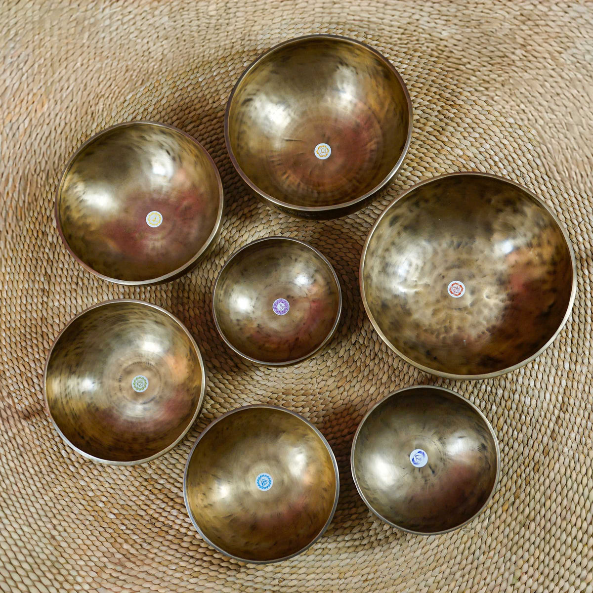 Seven handcrafted Himalayan singing bowls arranged together, featuring graduated sizes, chakra symbols, and warm antique bronze-gold finishes.