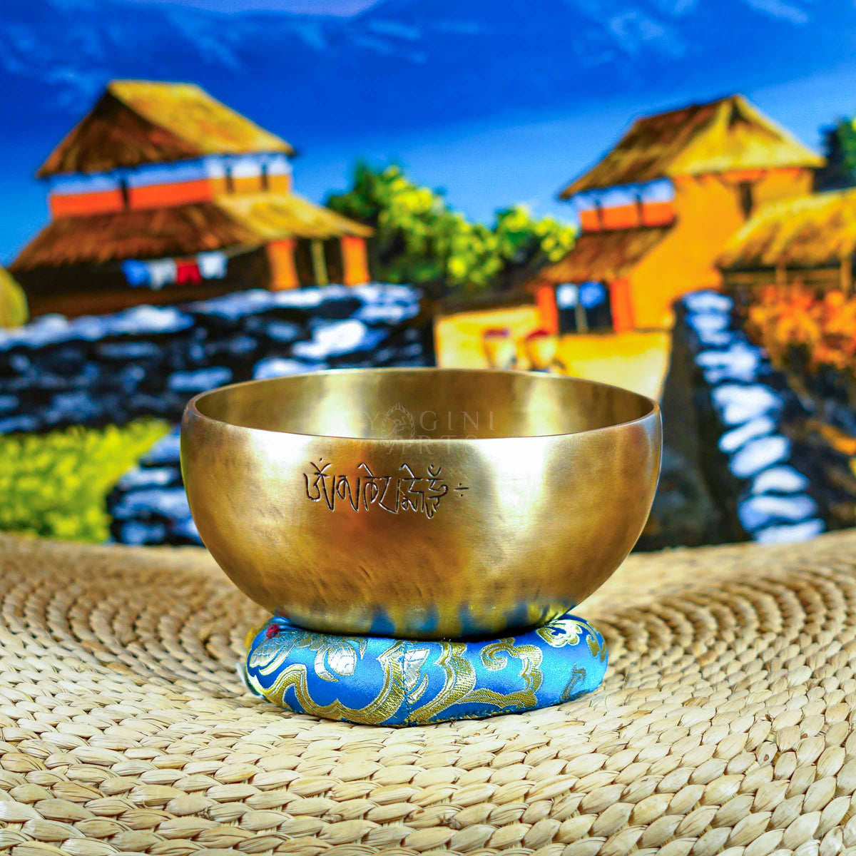 Front view of a handcrafted singing bowl with Om symbol, warm bronze finish, blue patterned cushion, and traditional wooden mallet.