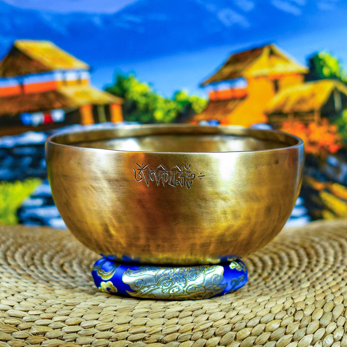 Front view of a handcrafted singing bowl with Om symbol, warm bronze finish, blue patterned cushion, and traditional wooden mallet.