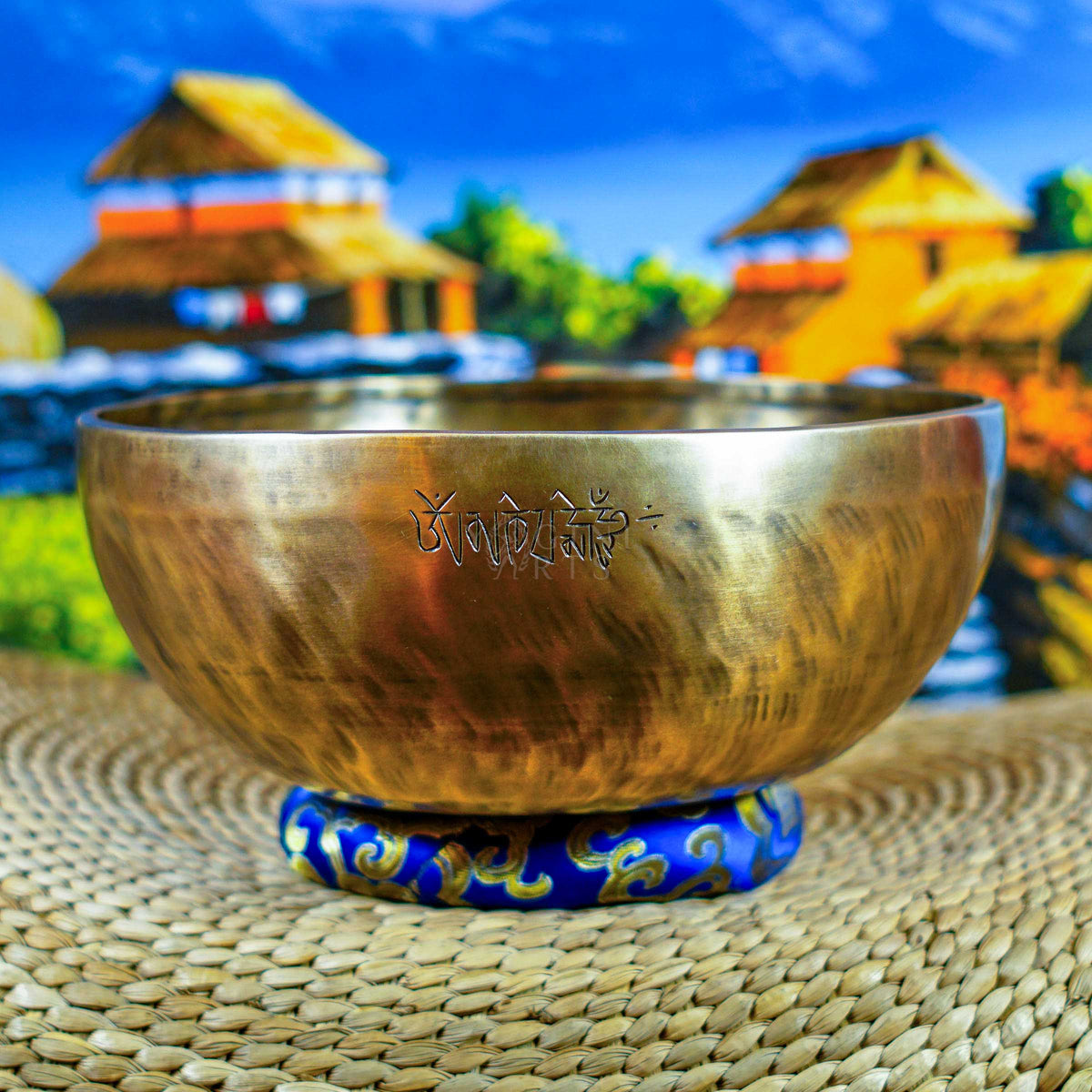 Front view of a handcrafted singing bowl with Om symbol, warm bronze finish, blue patterned cushion, and traditional wooden mallet.
