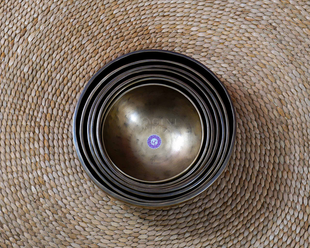 Seven handcrafted Himalayan singing bowls arranged together, featuring graduated sizes, chakra symbols, and warm antique bronze-gold finishes.