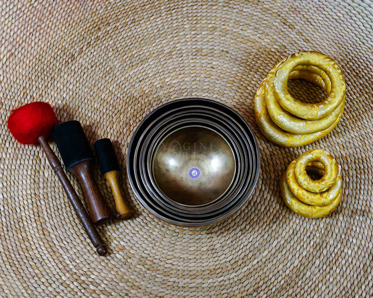 Seven handcrafted Himalayan singing bowls arranged together, featuring graduated sizes, chakra symbols, and warm antique bronze-gold finishes.