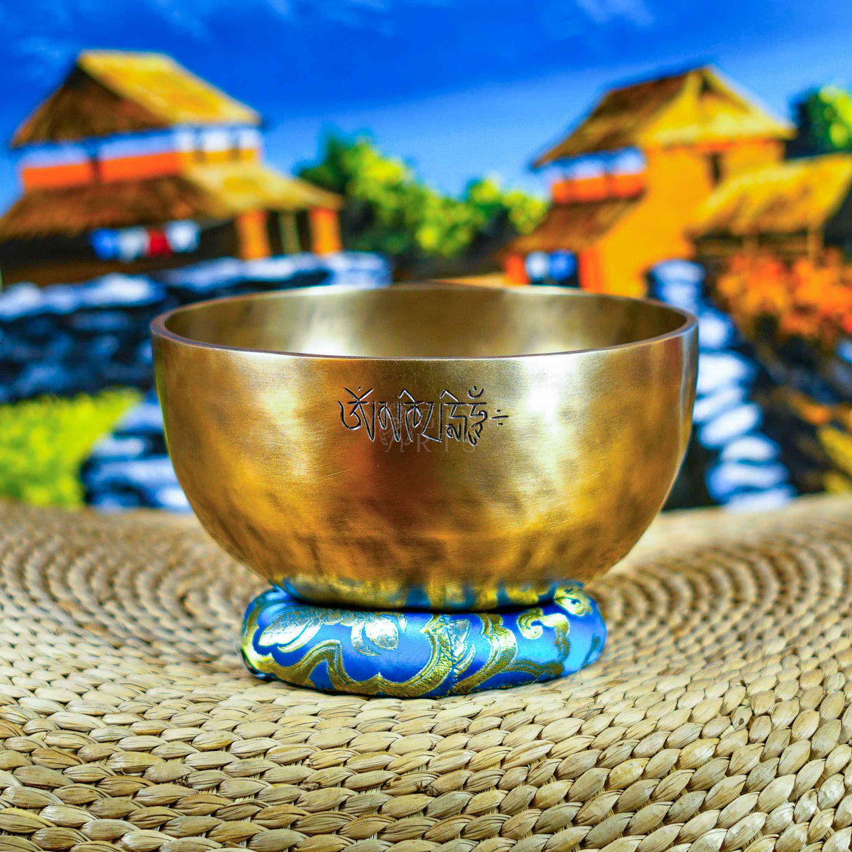 Front view of a handcrafted singing bowl with Om symbol, warm bronze finish, blue patterned cushion, and traditional wooden mallet.