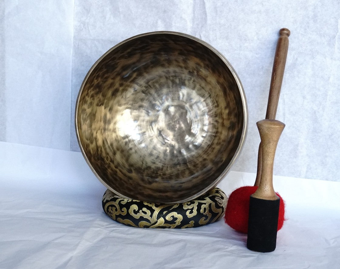 Hand-hammered brass singing bowl with a wooden striker and a red-padded mallet, displayed on a decorative cushion against a white background.