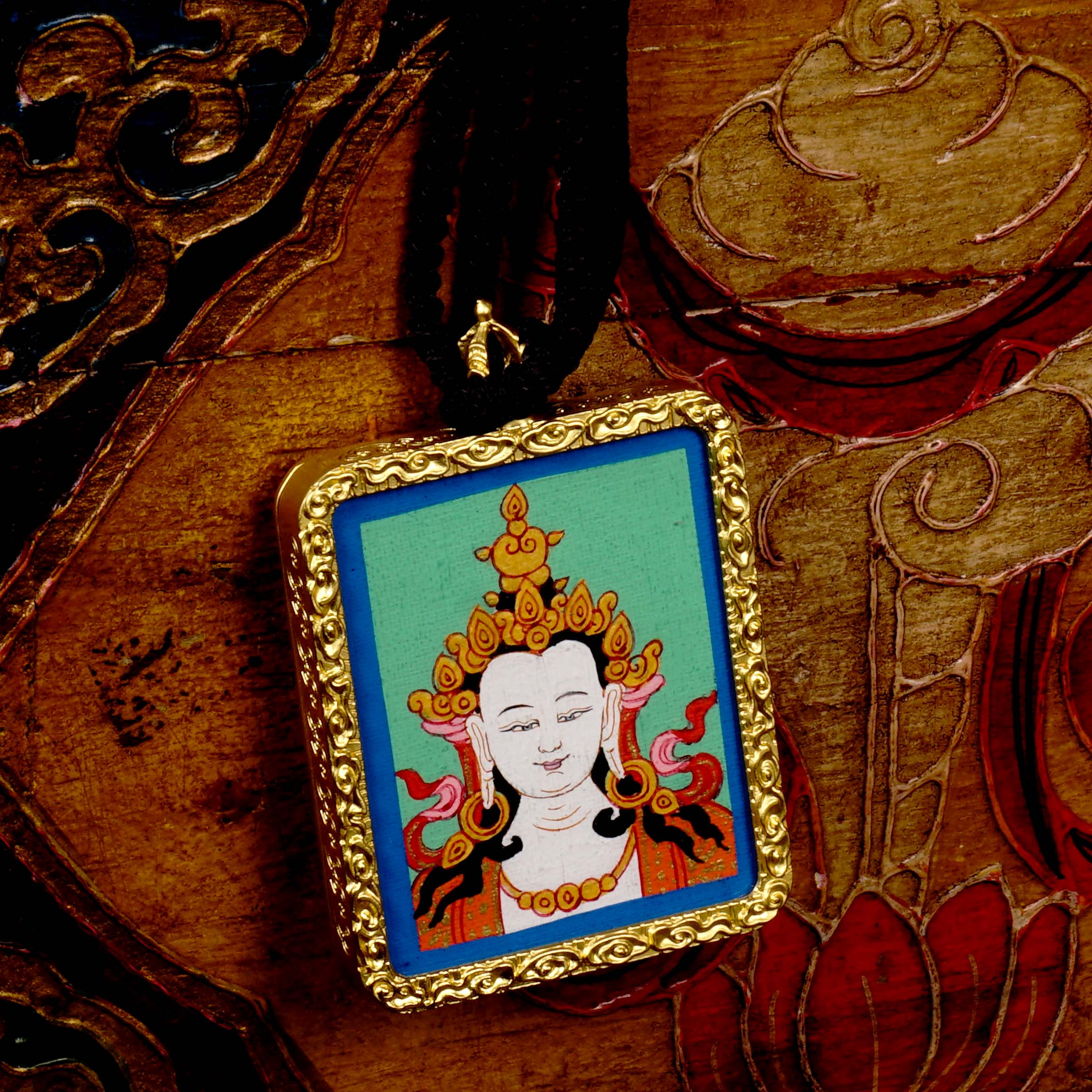 Square gold-framed Vajrasattva pendant with blue background, held in an open hand against a black backdrop.