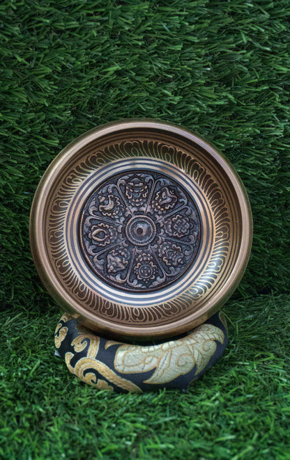 Top-down view of a bronze Tibetan singing bowl featuring an engraved Ashtamangala (Eight Auspicious Symbols) mandala design on its interior, resting on a black and gold silk cushion over green grass.
