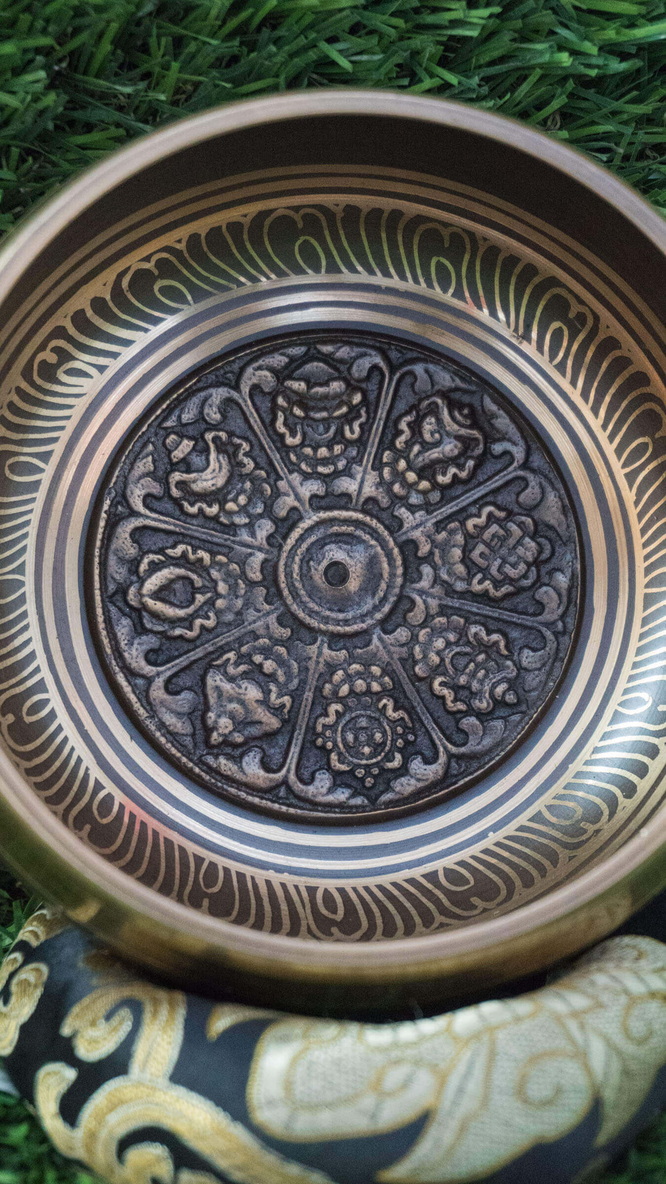 Top-down view of a bronze Tibetan singing bowl featuring an engraved Ashtamangala (Eight Auspicious Symbols) mandala design on its interior, resting on a black and gold silk cushion over green grass.