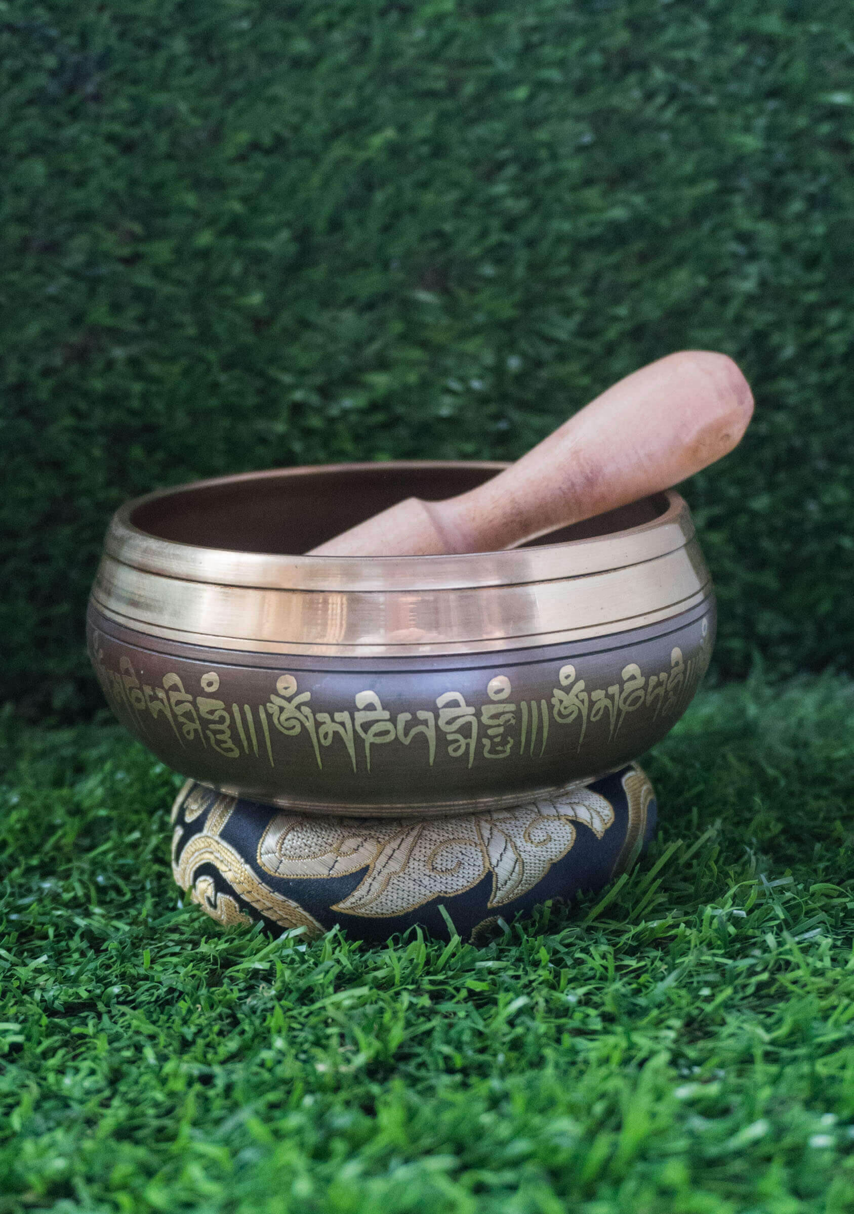 Top-down view of a bronze Tibetan singing bowl featuring an engraved Ashtamangala (Eight Auspicious Symbols) mandala design on its interior, resting on a black and gold silk cushion over green grass.