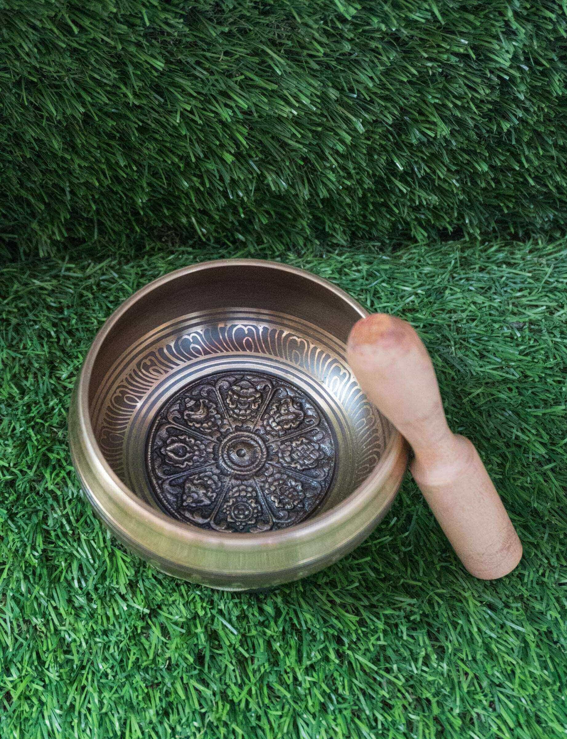 Top-down view of a bronze Tibetan singing bowl featuring an engraved Ashtamangala (Eight Auspicious Symbols) mandala design on its interior, resting on a black and gold silk cushion over green grass.