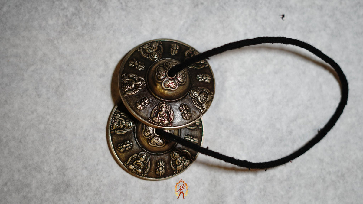 Close-up of bronze tingsha meditation cymbals with detailed Buddhist engravings, resting on a gray textured surface.
