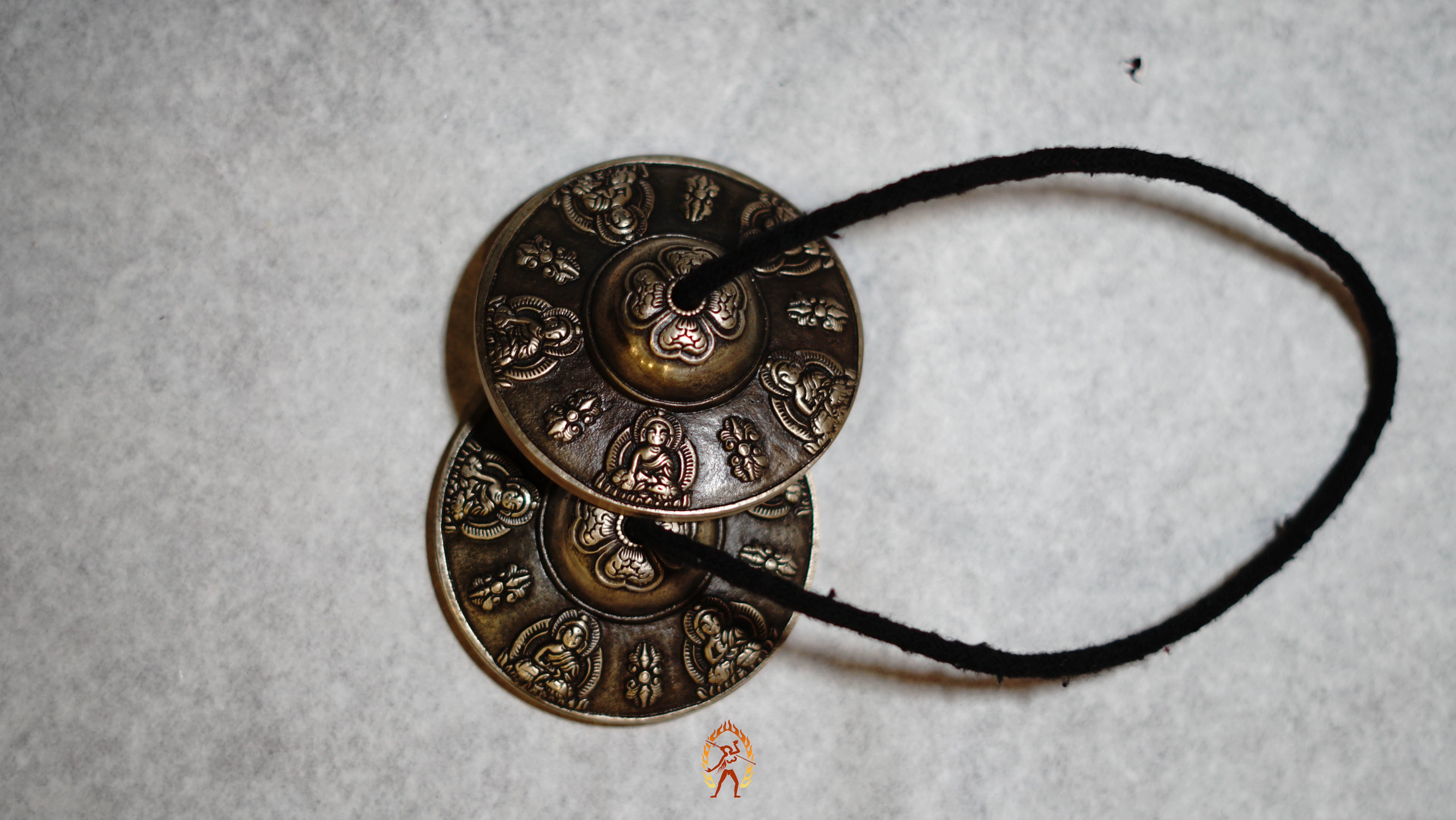 Close-up of bronze tingsha meditation cymbals with detailed Buddhist engravings, resting on a gray textured surface.