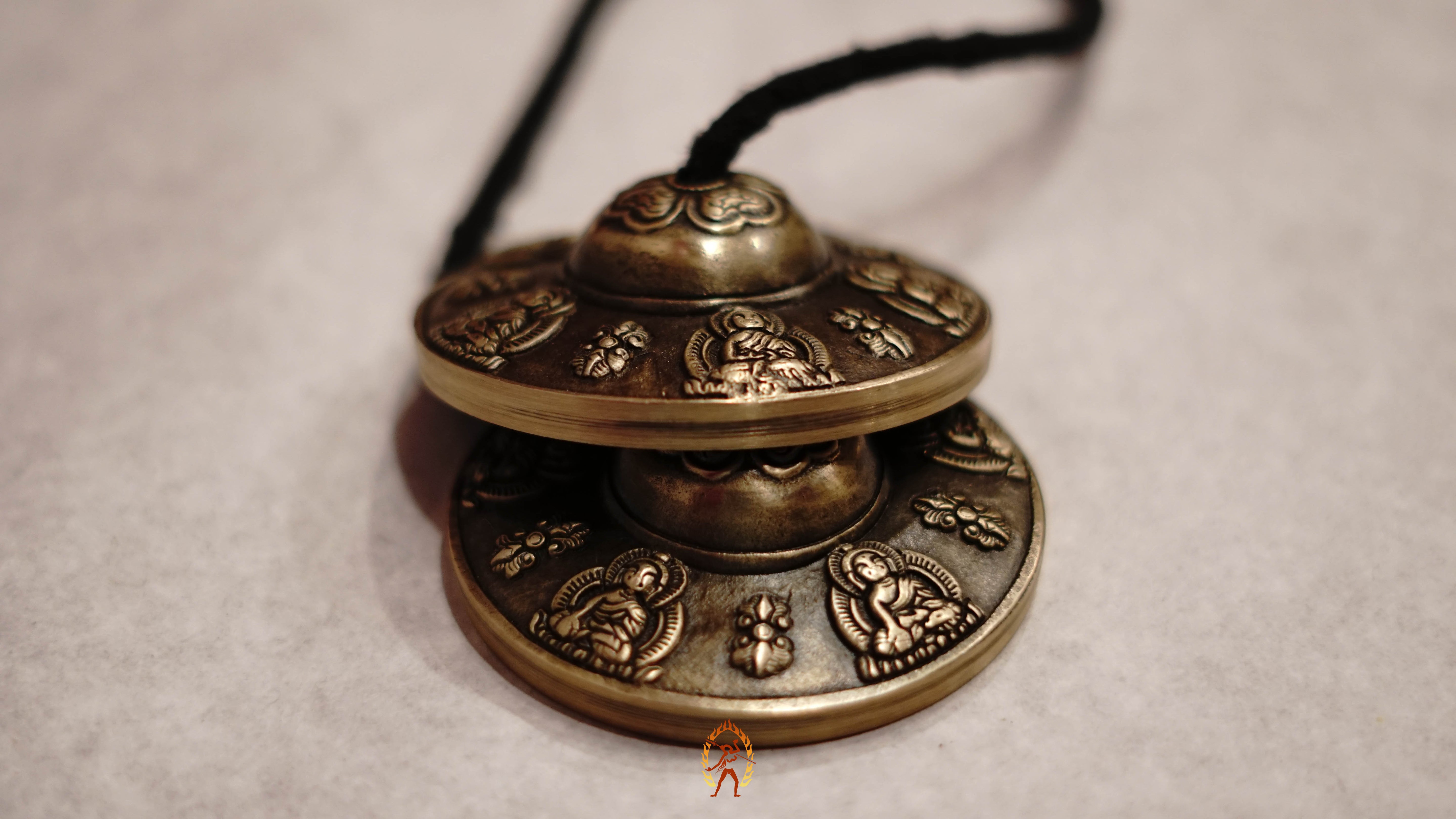 Close-up of bronze tingsha meditation cymbals with detailed Buddhist engravings, resting on a gray textured surface.