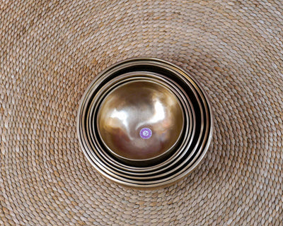 Seven handcrafted singing bowls nested together, showing their graduated sizes, smooth metal surfaces, and warm bronze-gold finish.