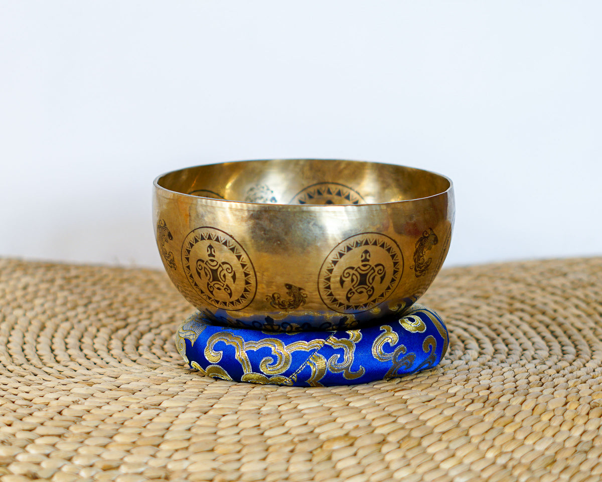 Golden Turtle Design singing bowl with repeating engraved motifs displayed on a blue brocade cushion.