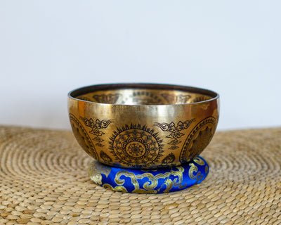 Golden floral mandala singing bowl with detailed ornamental engravings displayed on a blue brocade cushion.
